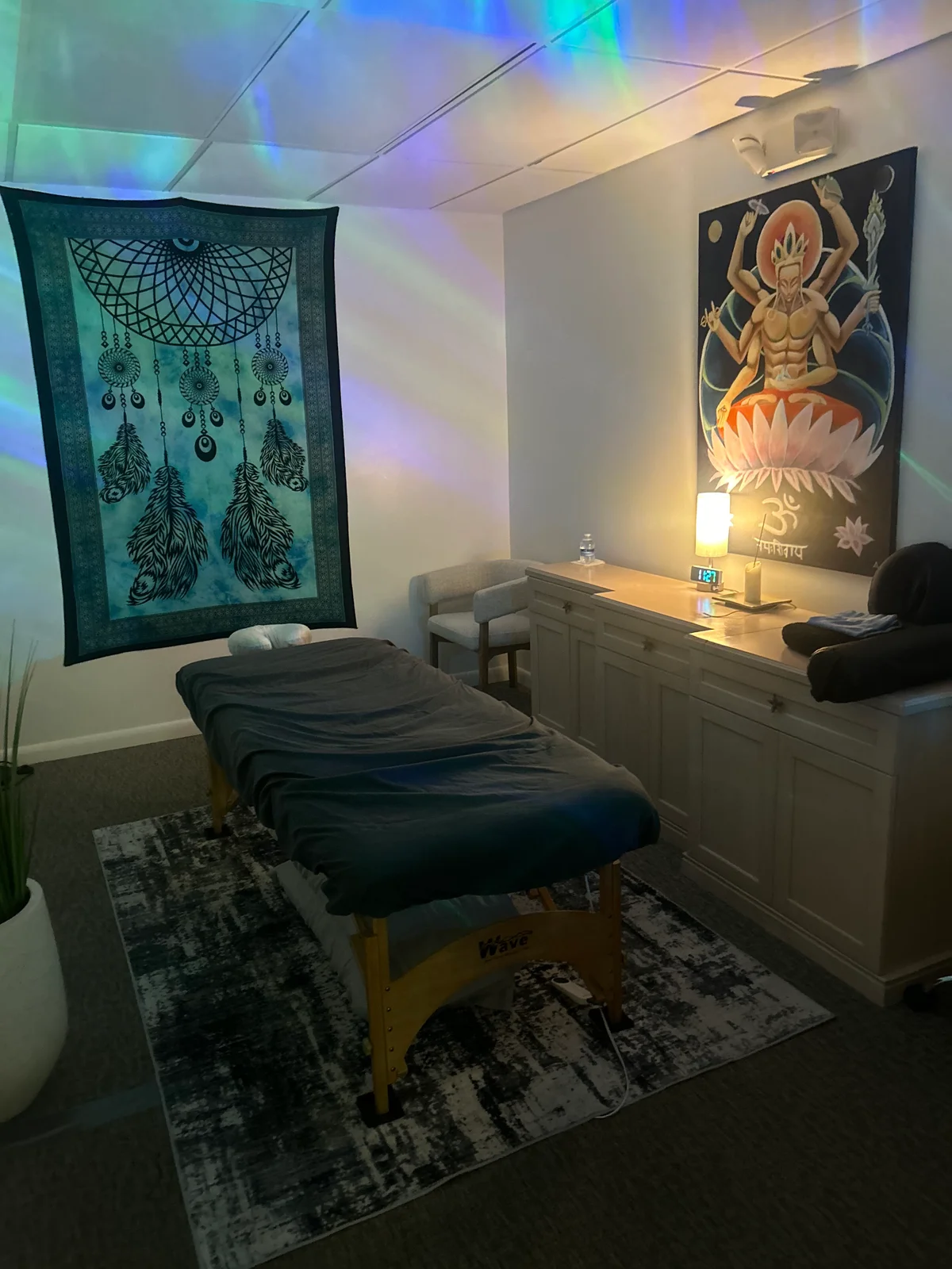 The massage room with blue and green ambient lighting, a dark-covered table, and a softly lit lamp